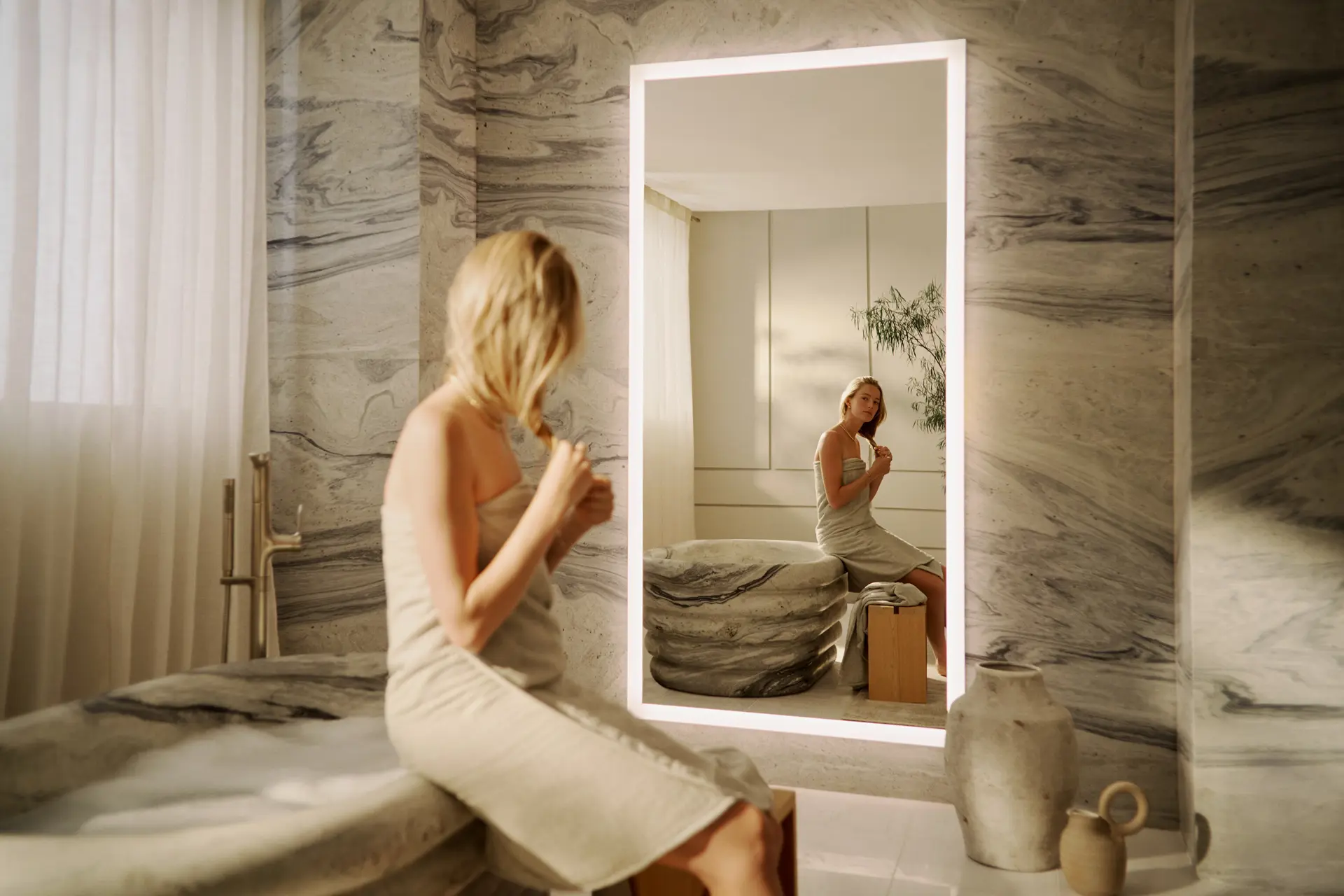 Woman in a wrapped towel seated before a backlit mirror in a veined marble bathroom