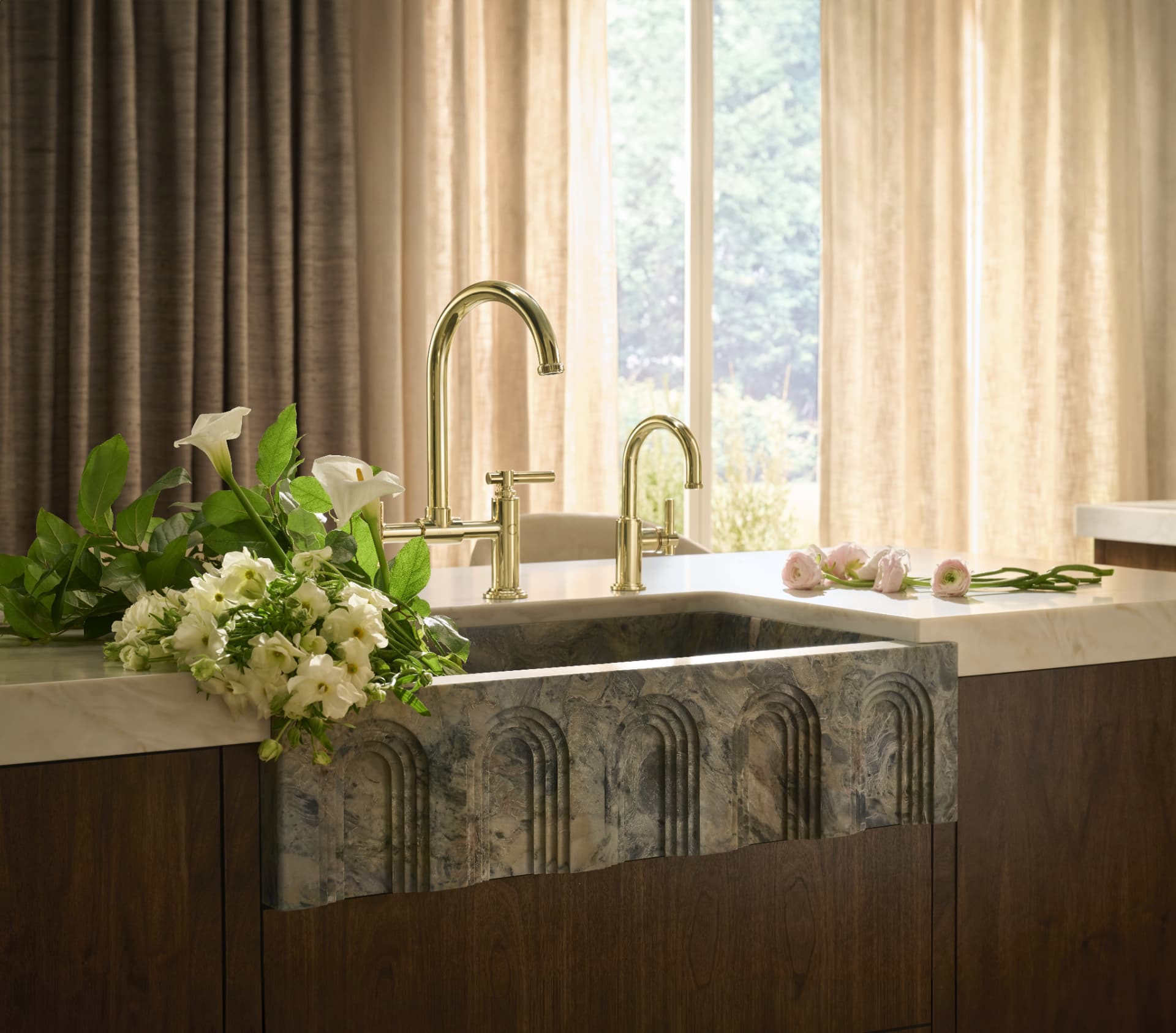 Carved stone farmhouse sink with brass bridge faucet and white blooms